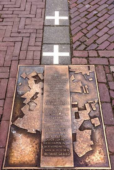 A bronze plaque embedded in a sidewalk, featuring a map-like design and engraved text, with white crosses marked on the pavement nearby.