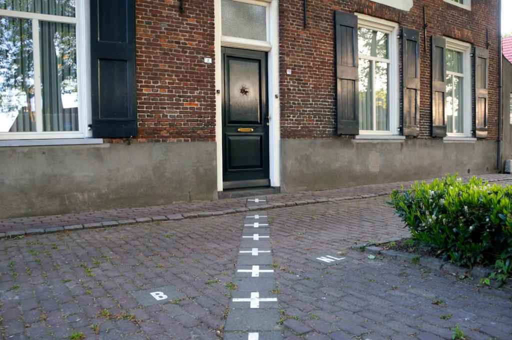 A cobblestone pathway leading to a dark green front door, bordered by a brick wall and windows with black shutters. White markings resembling a hopscotch grid are visible on the ground.